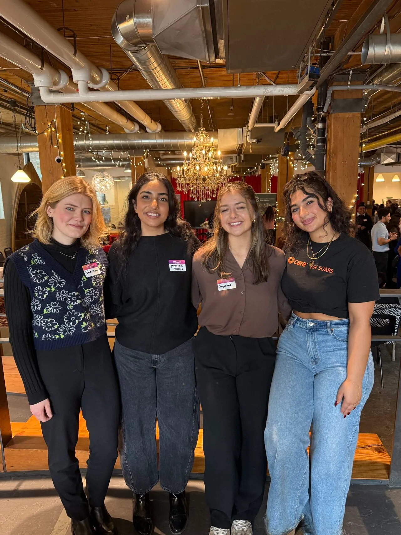 Four young women smiling together at a community event; one wears a CARE SHE SOARS t-shirt.