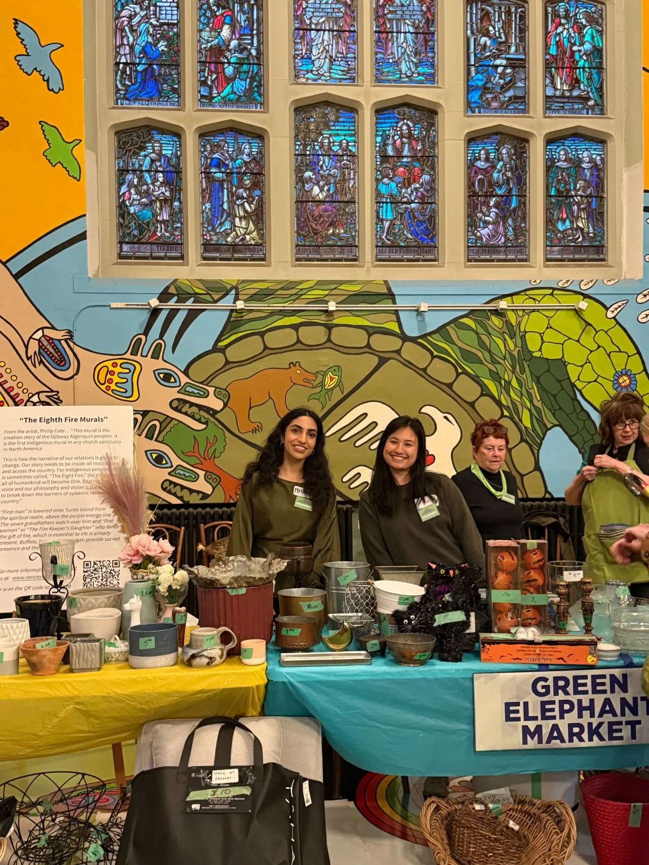 Two people smiling behind a table of second-hand goods at the Green Elephant Market, beneath stained glass windows and a large Indigenous mural.