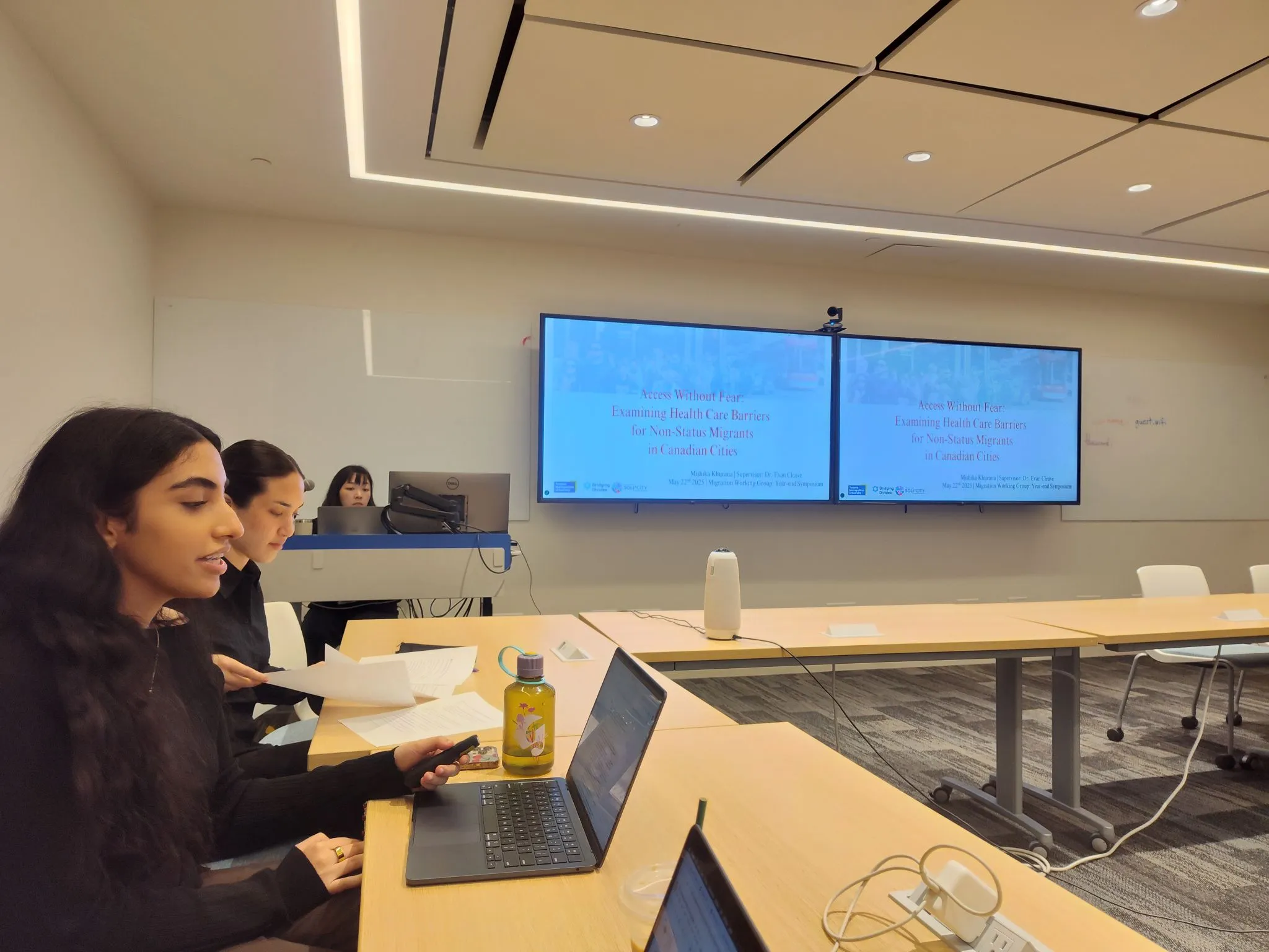 Mishika Khurana presenting at a boardroom table, with her research title slide, Access Without Fear, on two screens behind her.